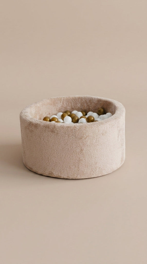 Ronde beige ballenbak van teddystof met witte, goudkleurige en lichtgrijze ballen op een beige achtergrond.