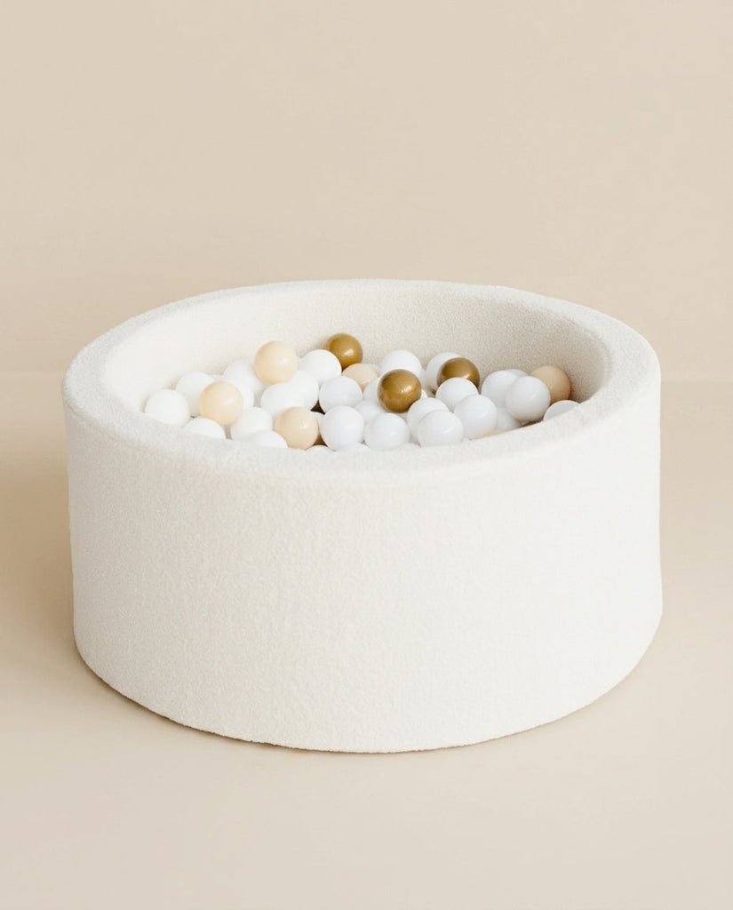 Rond, wit bouclé ballenbad met witte, beige en goudkleurige ballen tegen een beige achtergrond.
