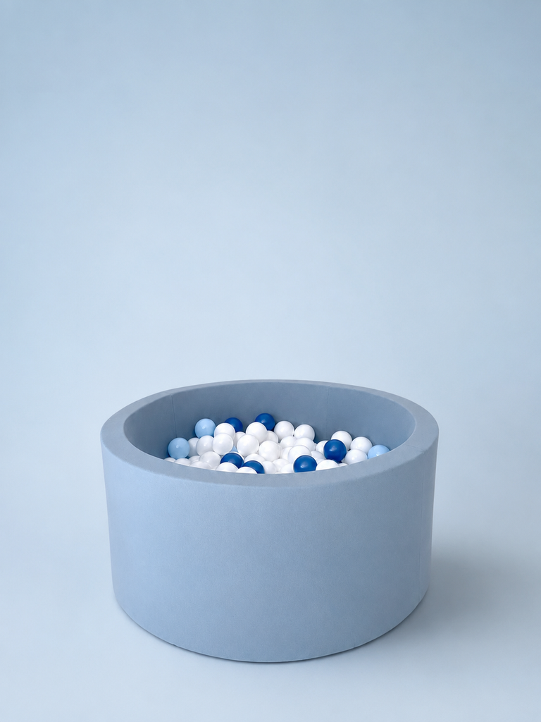 Ronde lichtblauwe stoffen ballenbak met witte, lichtblauwe en donkerblauwe ballen op een lichtblauwe achtergrond.