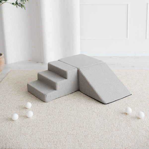 Grijze bouclé speelset met trapje, blok en helling op een licht vloerkleed met witte ballen.