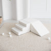Witte bouclé speelset met trapje, blok en driehoekige helling, schuin van boven gezien op een beige vloerkleed.