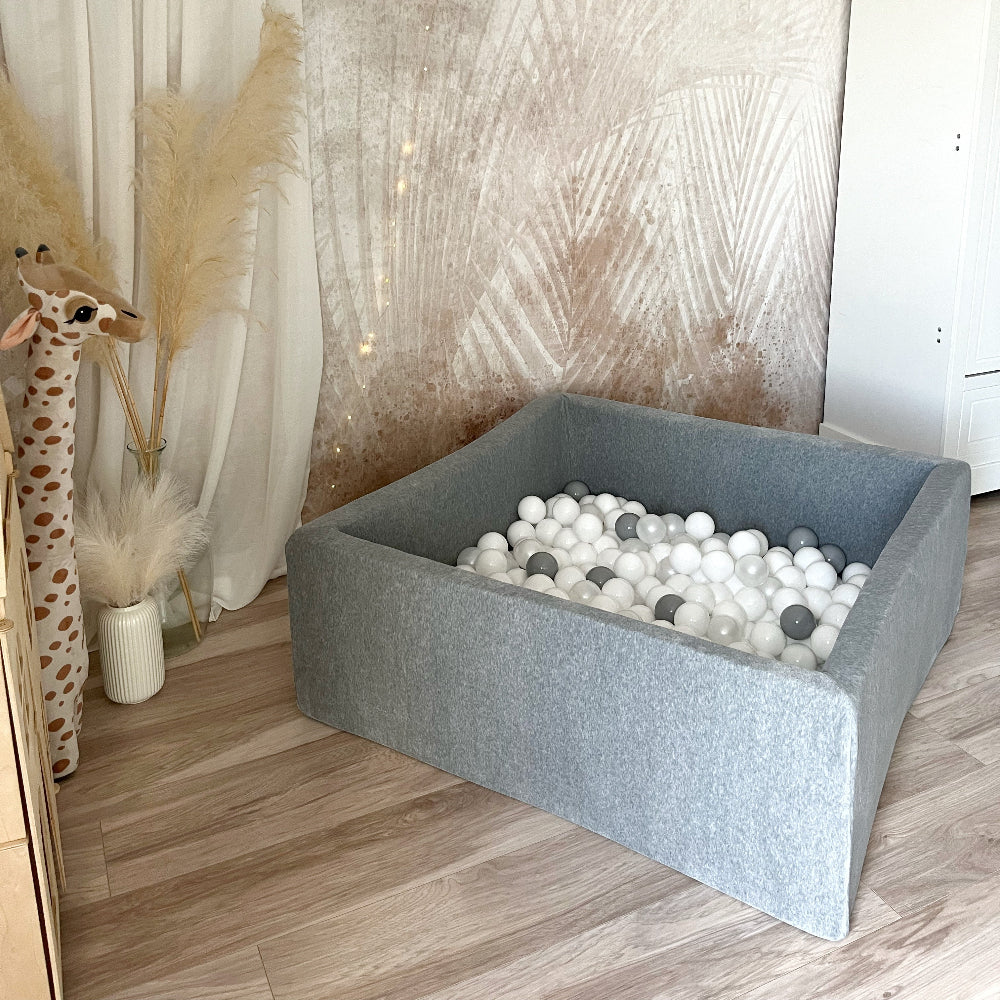Vierkant grijs velours ballenbad met witte en grijze balletjes op een houten vloer in een kinderkamer.