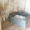 Vierkant grijs velours ballenbad met witte en grijze balletjes op een houten vloer in een kinderkamer.