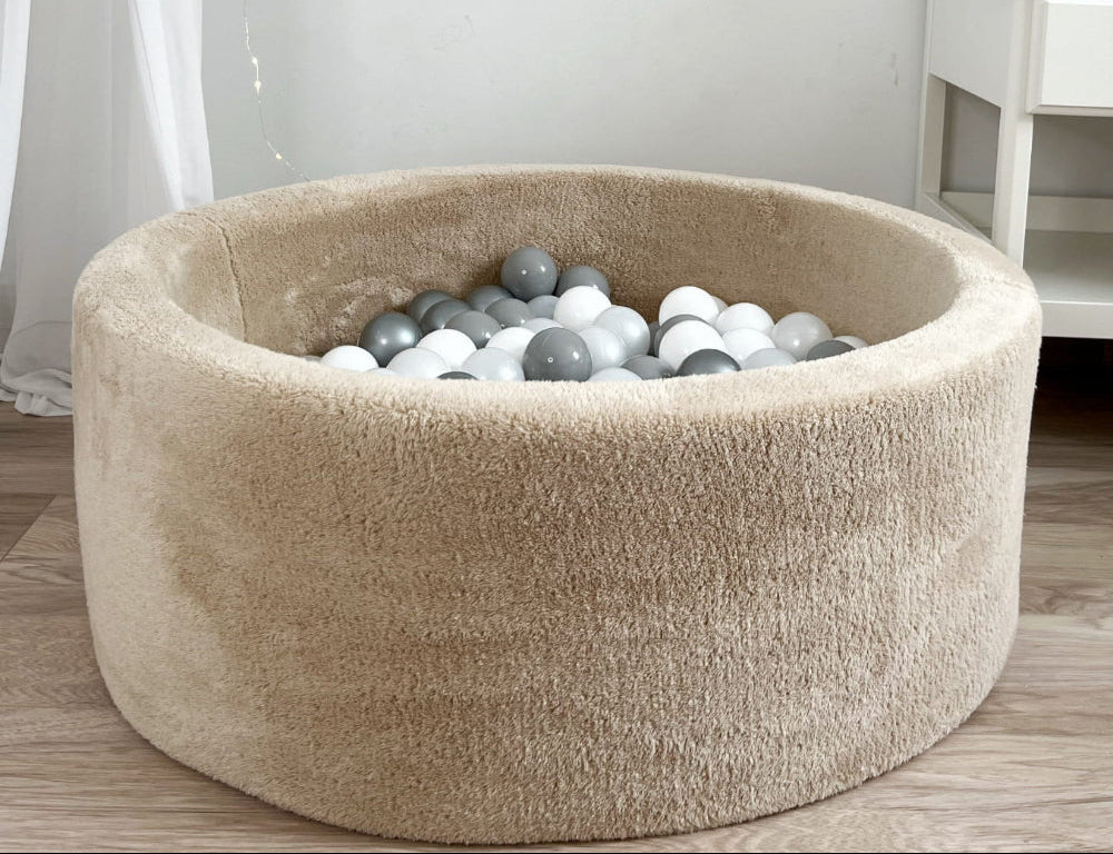 Rond beige teddy ballenbad met witte, lichtgrijze en donkergrijze balletjes op een houten vloer.