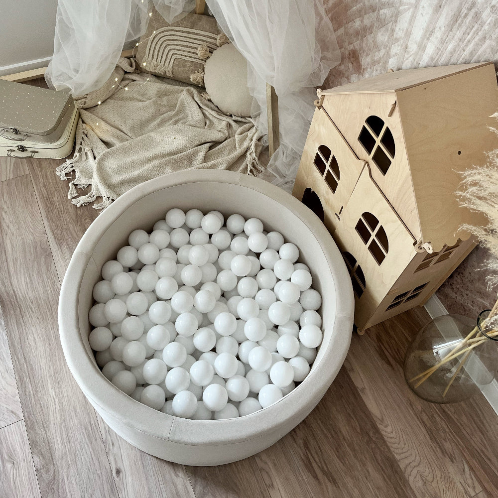 Bovenaanzicht van een rond crèmekleurig ballenbad gevuld met witte balletjes naast een houten poppenhuis.