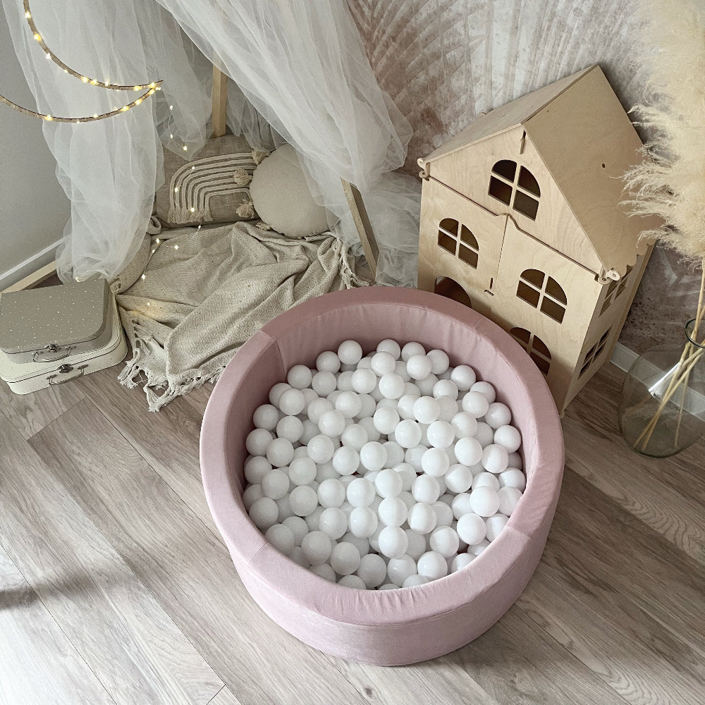 Lichtroze ballenbad met witte balletjes van bovenaf gezien, naast een houten speelhuisje en een speelhoek met klamboe.