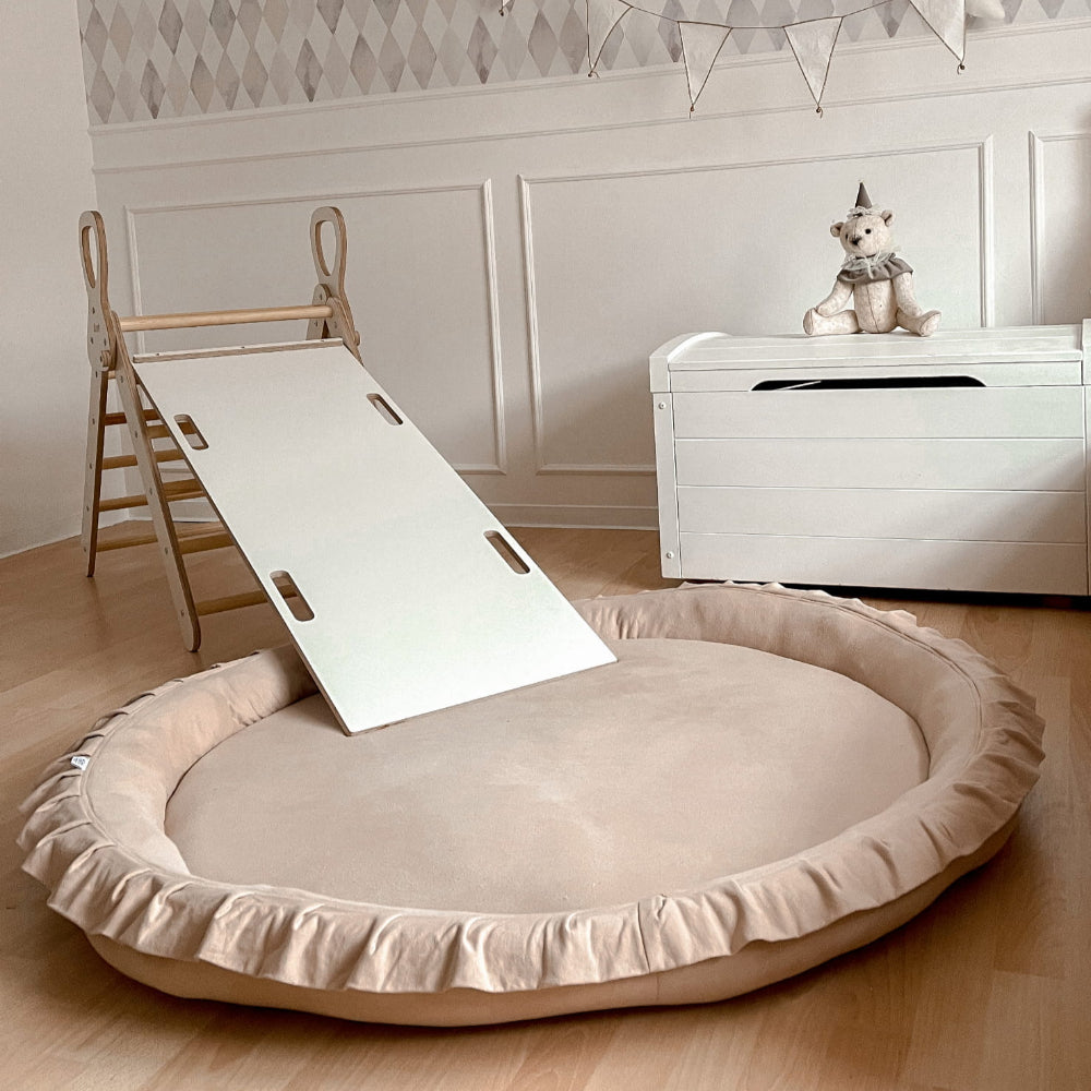 Beige speelnest met opstaande rand en ruches onder een glijbaan in een kinderkamer.